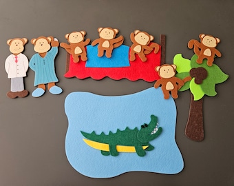 Könnte beinhalten: Filzspielzeug-Set mit einem braunen Alligator, einem braunen Baum mit grünen Blättern und sechs braunen Affen. Zwei der Affen befinden sich auf einem roten Bett mit blauer und weißer Bettwäsche. Zwei braune Figuren mit weißer und blauer Kleidung stehen links neben dem Bett.