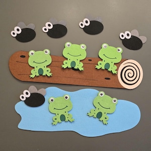 May include: A felt craft scene featuring green frogs and black flies. Three frogs sit on a brown log, while two frogs are on a blue water shape. Several black flies with grey wings are scattered around the scene.