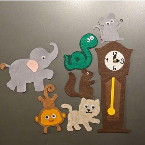 Op de afbeelding: Een set viltfiguren bestaande uit een grijze olifant, een groene slang, een bruine eekhoorn, een beige aap, een lichtbeige kat, een grijze muis en een bruine staande klok met een witte wijzerplaat, zwarte cijfers en een gele slinger.
