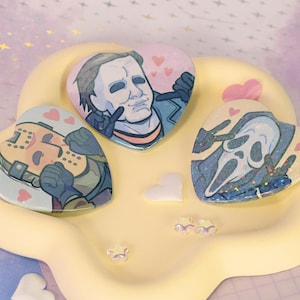 Op de afbeelding: Drie hartvormige pins met horrorfilmkarakters, rustend op een gele, wolkvormige tray. De pins tonen Michael Myers, Jason Voorhees en Ghostface. Roze en witte hartvormige decoraties zijn verspreid.