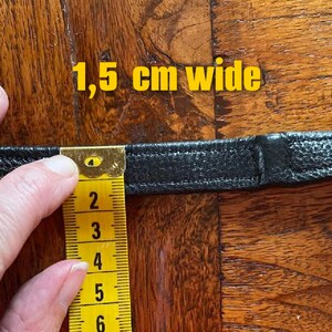 May include: Close-up of a black leather strap measured with a yellow tape measure. The tape measure reads 1.5 cm wide.