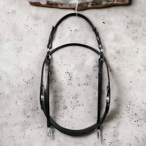 May include: Black leather horse bridle hanging on a white wall. The bridle has silver buckles and hardware.