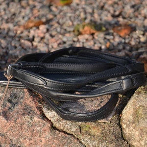 May include: A black leather horse riding bridle folded on a rocky surface. The bridle is made of high-quality leather and has a simple design.