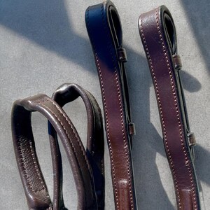 May include: Two brown leather reins with stitching detail. The reins are folded and laid out on a gray surface.