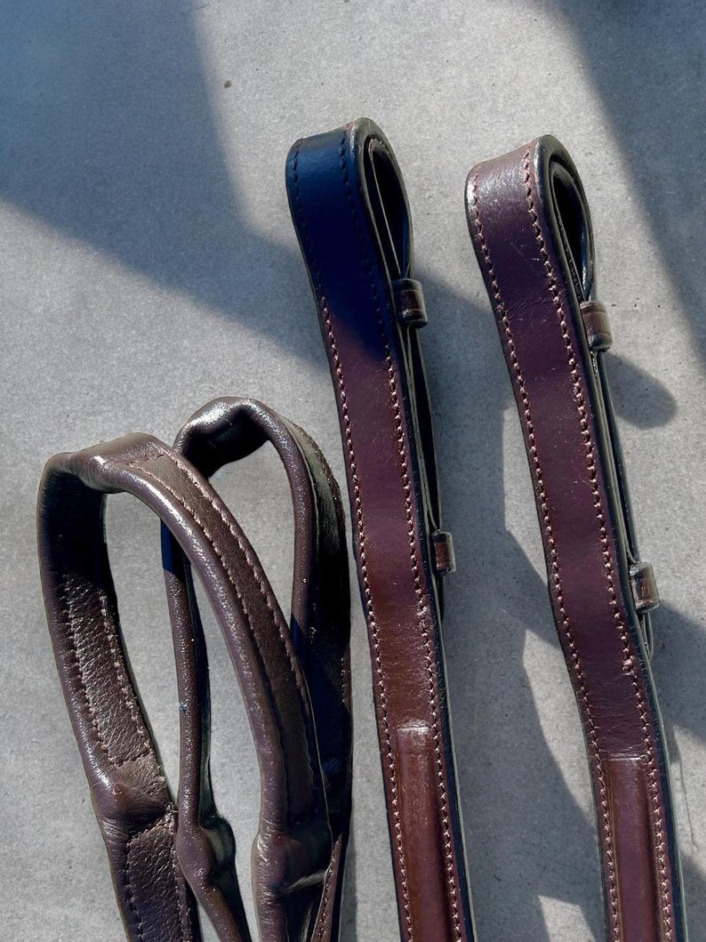 May include: Two brown leather reins with stitching detail. The reins are folded and laid out on a gray surface.