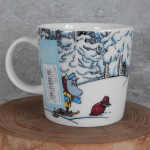 Könnte beinhalten: Weiße Keramiktasse mit einer Winterszene mit einer blauen und gelben Figur auf Skiern und einer roten Figur in einem verschneiten Wald. Die Tasse hat einen schwarzen Henkel und ein blaues und weißes Etikett mit dem Text "2016" und einem Barcode.