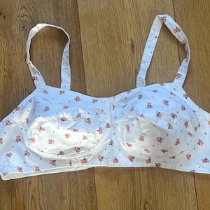 Op de afbeelding: Witte beha met verstelbare bandjes en een bloemenpatroon van kleine rode en roze bloemen. De beha heeft een klassiek ontwerp met beugelondersteuning en is gemaakt van een lichte stof.