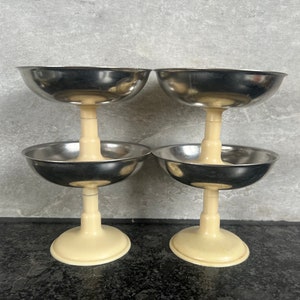 Peut inclure: Quatre coupelles à dessert en argent avec des bases crème empilées par paires. Les coupelles sont rondes et ont une surface lisse et réfléchissante.