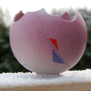 Könnte beinhalten: Eine kleine, rosafarbene Keramikvase mit abgebrochenem Rand und einem geometrischen Muster in Blau und Rot. Die Vase steht auf einer schneebedeckten Oberfläche.