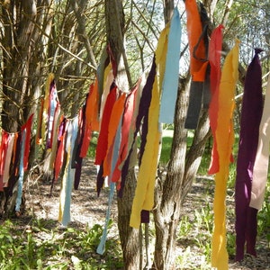 May include: A colorful fabric garland hangs from tree branches in a forest setting. The garland is made of various colors and textures of fabric, including red, orange, yellow, green, blue, purple, and white.