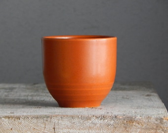 May include: A small, orange ceramic cup with a rounded shape. The cup has a smooth, matte finish and is set against a neutral background. The cup is likely used for drinking beverages.