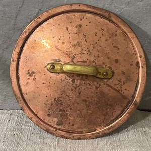 Könnte beinhalten: Ein runder Kupfertopfdeckel mit einem Messinggriff. Der Deckel hat ein leicht abgenutztes Aussehen mit dunklen Flecken und einem sichtbaren Rand. Der Griff ist mit zwei Nieten an jedem Ende befestigt.