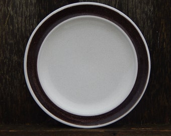 May include: A white ceramic plate with a brown rim. The plate is round and has a smooth surface.