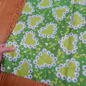 May include: Green fabric with a repeating pattern of lime green hearts outlined with white daisies and blue centers. Small green hearts are scattered throughout the design. The fabric is being held by a hand.