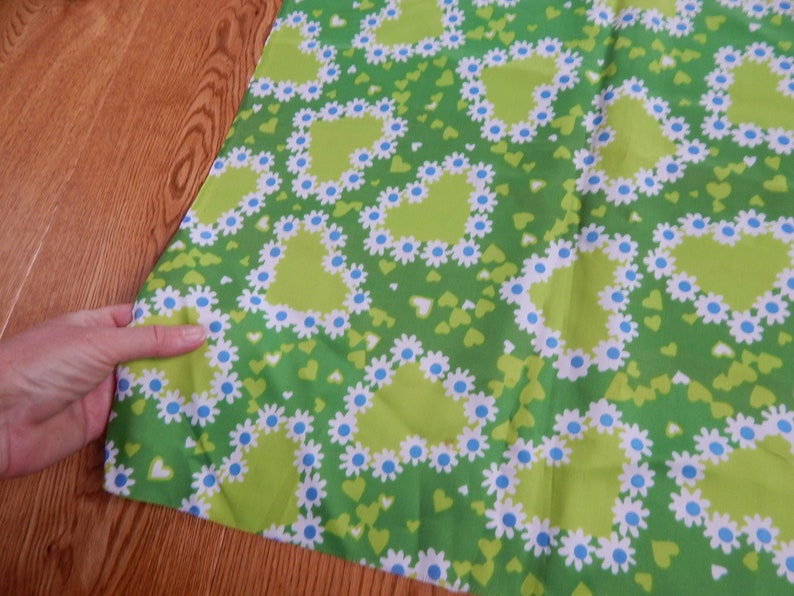 May include: Green fabric with a repeating pattern of lime green hearts outlined with white daisies and blue centers. Small green hearts are scattered throughout the design. The fabric is being held by a hand.