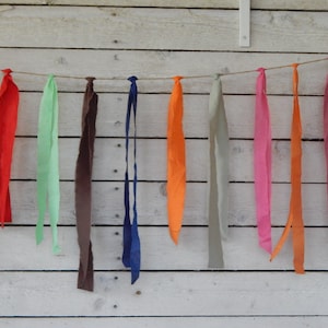 May include: A string of fabric scraps in various colors, including yellow, red, green, brown, blue, orange, pink, and gray, is strung on a twine line against a white wood background.