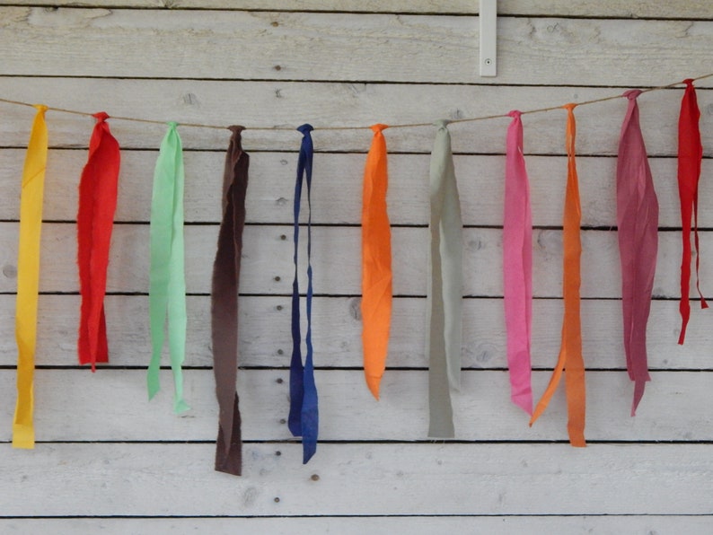 May include: A string of fabric scraps in various colors, including yellow, red, green, brown, blue, orange, pink, and gray, is strung on a twine line against a white wood background.