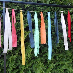 May include: A string of colorful fabric strips hanging from a metal fence. The strips are various shades of pink, white, red, yellow, blue, green, and orange.