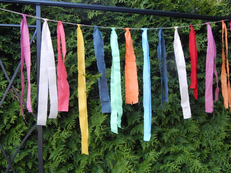 May include: A string of colorful fabric strips hanging from a metal fence. The strips are various shades of pink, white, red, yellow, blue, green, and orange.