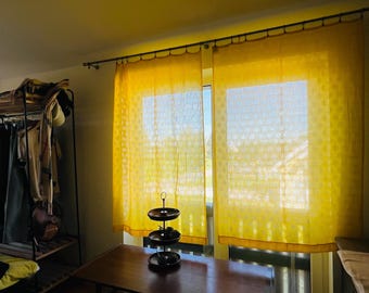 May include: Two sheer yellow curtains with a subtle circular pattern hang from a dark metal rod over a window. The fabric is translucent, allowing sunlight to filter through, and the curtains feature a simple hem at the bottom edge.