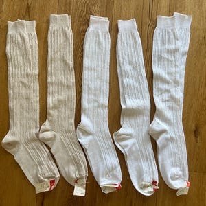 Könnte beinhalten: Fünf Paar kniehohe Socken. Zwei Paar sind beige, drei sind weiß. Alle haben eine gerippte Struktur. Die Socken liegen auf einer Holzoberfläche. Sie sind etwa 40 cm lang.