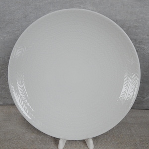 Puede incluir: Un plato de cena de cerámica blanca con una superficie texturizada y un sutil diseño de hojas en relieve alrededor del borde.