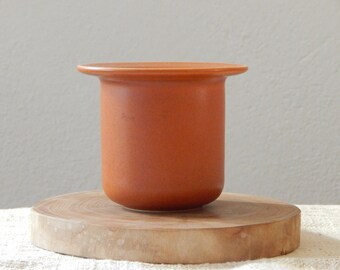 May include: A terracotta colored ceramic planter with a slightly flared rim. The planter is sitting on a wooden round slice.