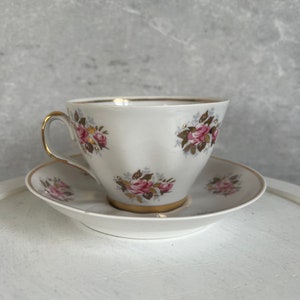 May include: A white teacup and saucer with a floral pattern of pink roses and gold accents. The cup has a gold rim and a handle.