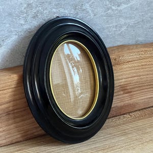 Può includere: Una cornice ovale nera con un bordo interno dorato. La cornice è realizzata in un materiale lucido e ha un design semplice ed elegante. La cornice è vuota, pronta per esporre una foto o un'opera d'arte.