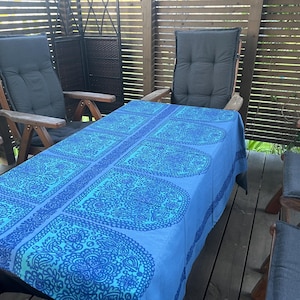 Puede incluir: Un mantel rectangular con un intrincado patrón floral y de cachemira en azul vibrante y azul oscuro. La tela cubre una mesa rodeada de sillas de madera con cojines gris oscuro, frente a una pantalla de privacidad de madera con listones.