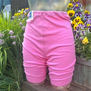 Peut inclure: Short rose avec un design froncé sur les côtés. Le short est fait d'un tissu doux et possède une taille élastique. Une petite étiquette est visible. Le short est présenté sur un mannequin devant un jardin.