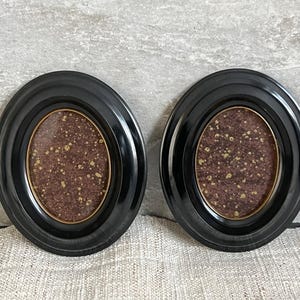 May include: Two black oval picture frames with a gold inner border. The frames have a dark brown and gold speckled background. The frames are displayed on a neutral-coloured surface.