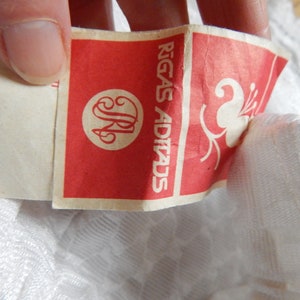 May include: A white fabric label with red text that reads "Rīgas Adītads" and a stylized flower design. The label has a circular logo with the letters "JR" inside.