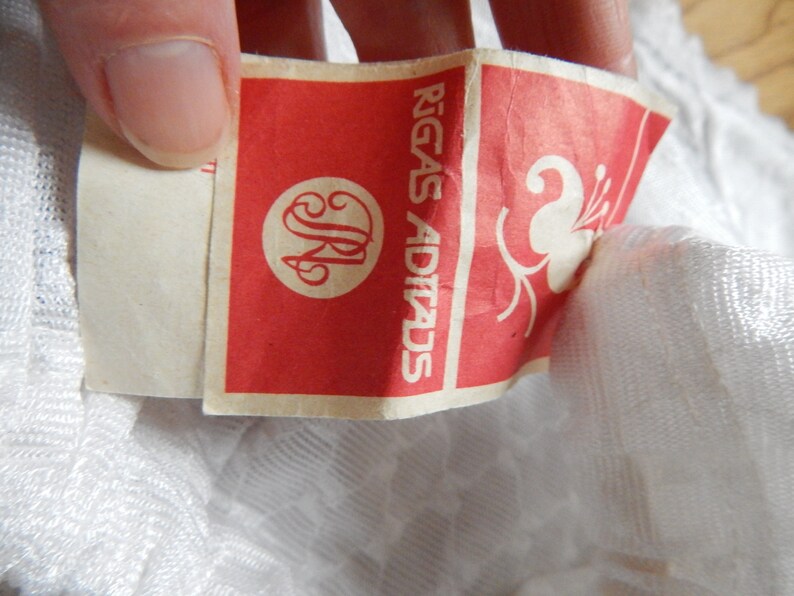 May include: A white fabric label with red text that reads "Rīgas Adītads" and a stylized flower design. The label has a circular logo with the letters "JR" inside.
