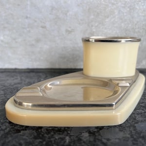 Op de afbeelding: Een vintage asbak van ivoor en zilver met een apart compartiment voor sigaretten. De asbak heeft een afgeronde rechthoekige vorm en een glad, gepolijst oppervlak.