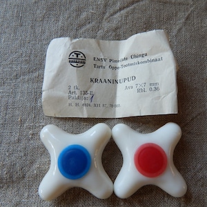 Könnte beinhalten: Zwei weiße Kunststoff-Wasserhahnknöpfe mit blauen und roten Mittelpunkten. Die Knöpfe sind mit "KRAANINUPUD" und dem Text "Ava 7x7 mm Rbl. 0.36" beschriftet.