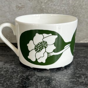 May include: A white ceramic teacup with a green floral design. The cup has a rounded handle and a wide rim.