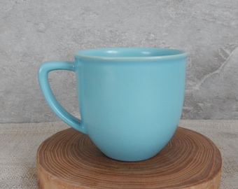 May include: A light blue ceramic mug with a rounded shape and a handle. The mug is sitting on a wooden surface.