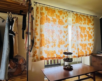 May include: A pair of sheer curtains featuring a repeating orange leaf pattern hanging on a metal rod, positioned in front of a window. A three-tiered dark brown serving stand sits on a wooden table in the foreground of the room.