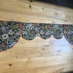 May include: A floral valance with a scalloped edge, featuring a dark background and a pattern of white, purple, and green flowers. The valance has a coral-coloured trim and is displayed against a wooden wall.