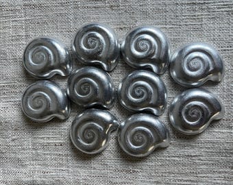 May include: A collection of ten silver-colored, shell-shaped decorative objects. Each shell has a spiral design and a small star-like emblem in the center. The objects are arranged on a textured, neutral-colored surface.