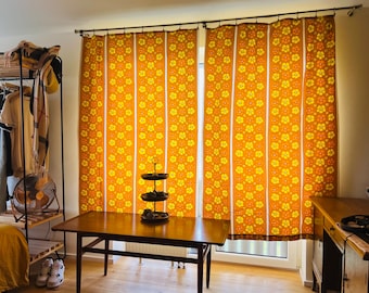 May include: A pair of floor-length curtains featuring a repeating pattern of orange and yellow flowers arranged in vertical stripes. The fabric is hung from a metal rod with rings, covering a window and displaying a vibrant floral design.