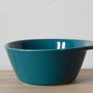 Puede incluir: Un tazón de cerámica de color verde azulado con forma redondeada y una pequeña asa en el lateral.