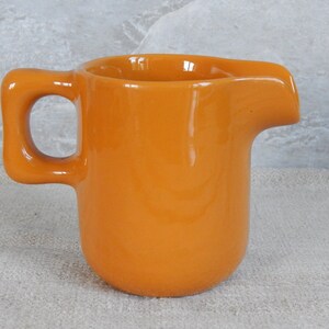 May include: A small, orange ceramic creamer with a rounded body and a handle. The creamer is ideal for serving milk or cream with coffee or tea.