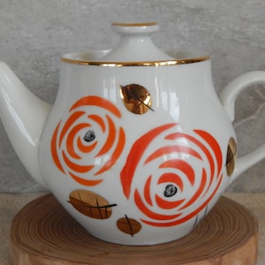 Könnte beinhalten: Weiße Teekanne mit goldener Verzierung und orangefarbenem Blumendesign. Die Teekanne hat einen Deckel und einen Henkel.
