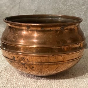 May include: A copper bowl with a rounded base and a wide, slightly flared rim. The bowl has horizontal bands around its body and shows signs of age and use. The copper has a warm, reddish-brown hue, suggesting it is an antique or vintage piece.