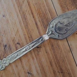 Op de afbeelding: Een zilveren serveerschep met een sierlijk ontwerp. De lepel heeft een decoratief handvat met een bloemenpatroon en een brede, platte schep.