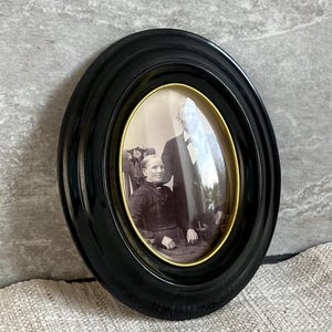May include: An oval, black framed photograph featuring a vintage portrait of a seated person and another standing behind them. The photograph is in black and white, with a gold-coloured inner frame.