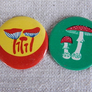May include: Two round pinback buttons with mushroom designs. The first button has a yellow background with red trim and three white mushrooms with red caps. The second button has a green background with two white mushrooms with red caps and white spots.