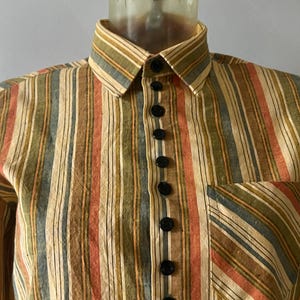 Può includere: Una camicia a maniche lunghe con bottoni e un motivo a righe in tonalità di marrone, arancione, verde e blu. La camicia ha un colletto classico e una chiusura con bottoni.